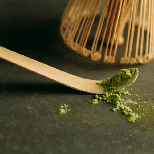 Bamboo chashaku scoop holding green matcha powder beside a traditional matcha whisk