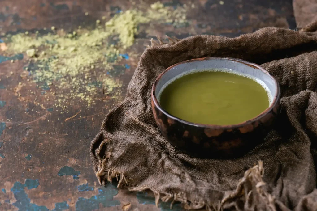Prepared matcha tea in a ceramic bowl on rustic cloth with matcha powder scattered on a wooden surface