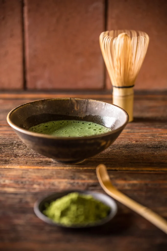 Traditional bowl of prepared matcha tea with bamboo whisk, scoop and matcha powder on a rustic wooden surface