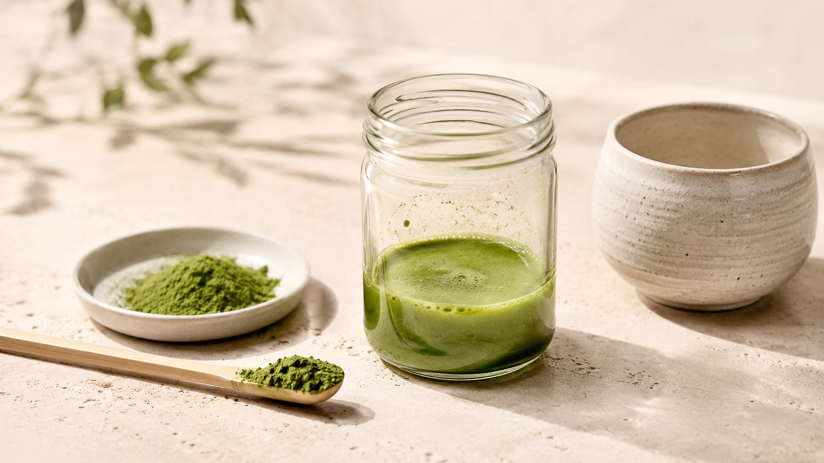 Clear glass jar with mixed green matcha beside a bamboo scoop, sifted matcha powder, and ceramic cup on a cream stone surface